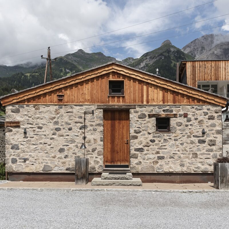 Alpine stone cottage with a stone lower wall and wooden upper façade, a wooden door, small window, and stone steps leading up.
