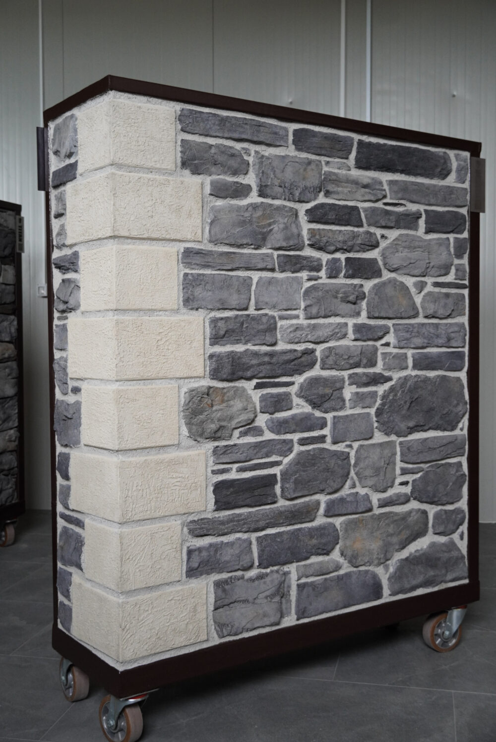 Display panel showing a faux stone wall sample with gray river stones and beige rectangular blocks on a wheeled stand in a showroom