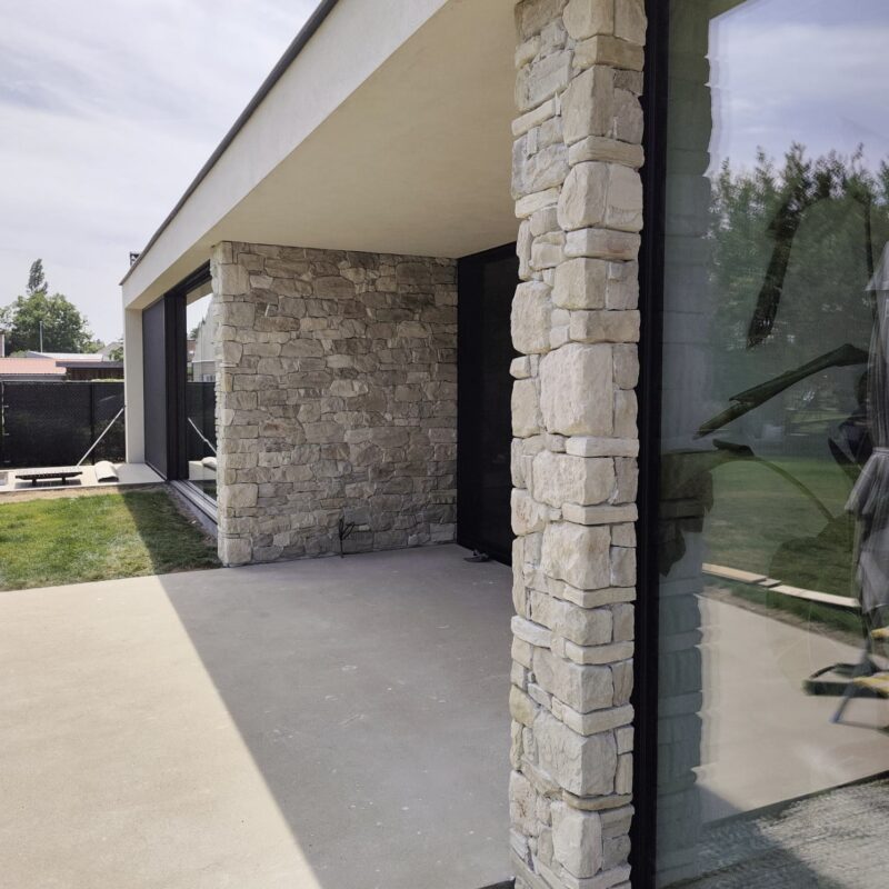 Modern house exterior featuring light stone veneer columns, large glass windows, and a concrete driveway under an overhang.