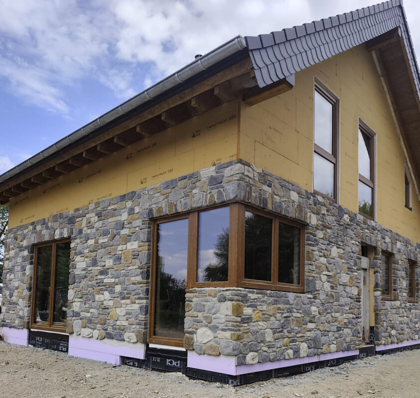 Under-construction house with stone veneer on the lower floor, yellow exterior sheathing on the upper level, and large wooden-framed windows under a tiled roof
